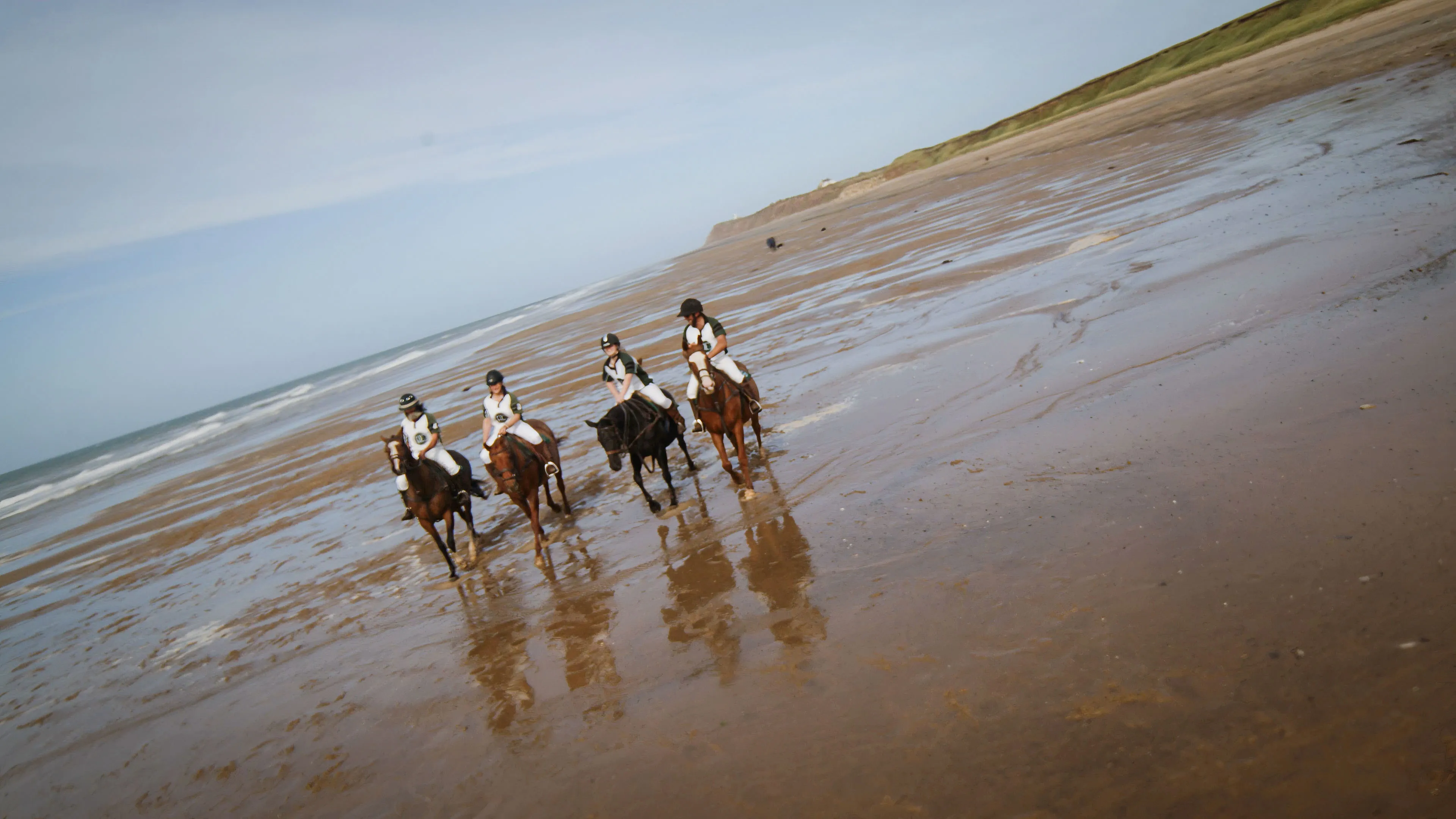 Four people ride horses along a wide, reflective beach with soft waves in the background and grassy dunes on the right, evoking a serene, adventurous mood.